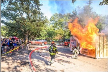 Image is of a group of people watching the Fire Department trying to put out a simulated dorm fire.