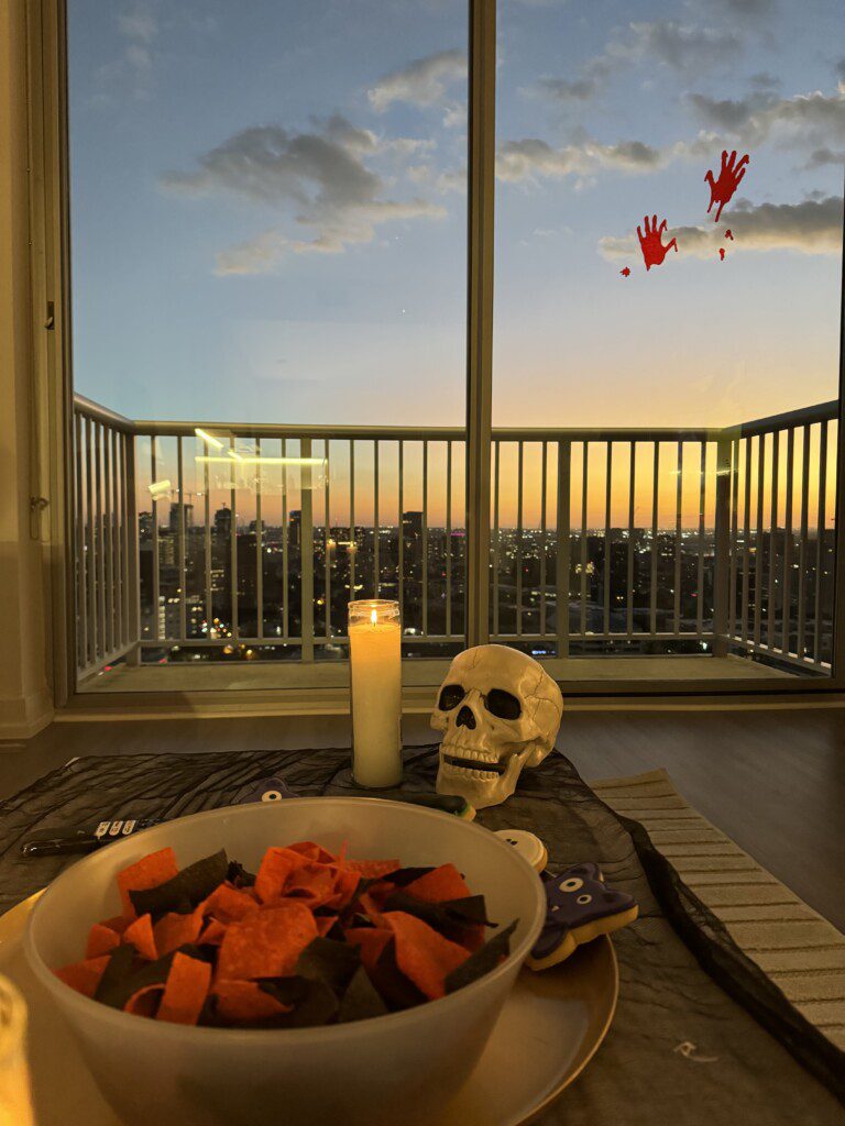 Photo of a skeleton bowl with halloween candy inside in front of a sliding glass door. In the background, there are bloody handprints on the sliding glass door.