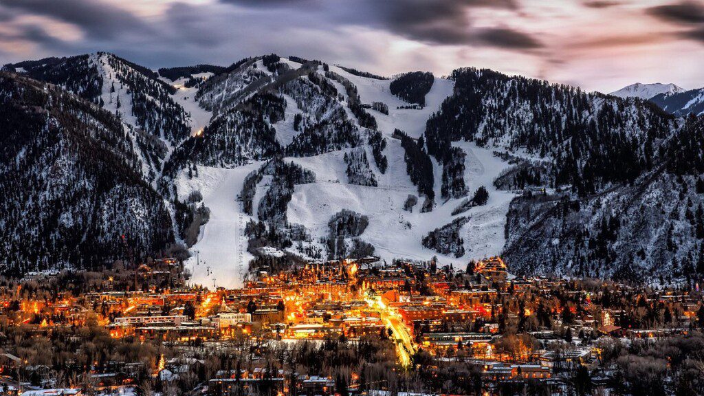 Image is of Aspen, Colorado lit up in the fore front with mountains with some snow in the background.