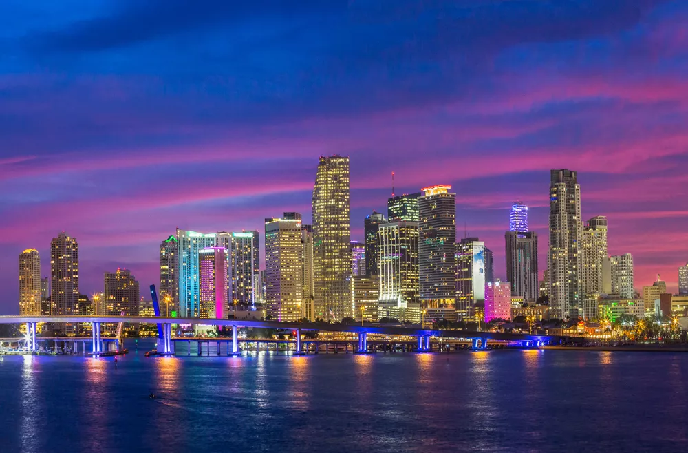 Image is of the Miami skyline at night with the bridge lit up in purple and orange.