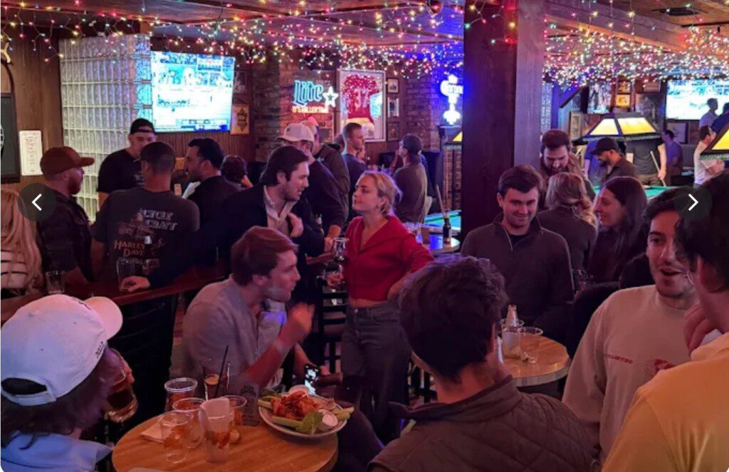 A bar with twinkle lights hanging from the ceiling. Contains a large crowd of college students enjoying each others company